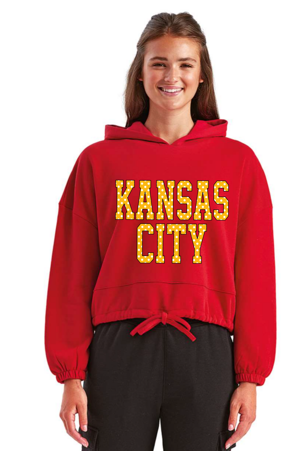 A woman modeling a red cropped hooded sweatshirt with 'KANSAS CITY' printed in yellow across the chest, paired with black sweatpants, against a plain white background.