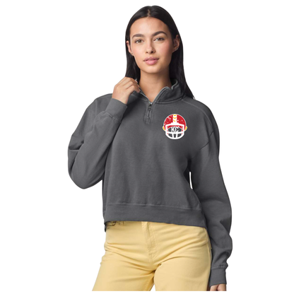 A woman modeling a dark gray cropped quarter-zip sweatshirt with a red, white, and yellow football helmet graphic on the left chest, paired with light yellow pants, against a plain white background.