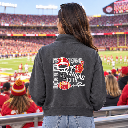 A woman viewed from behind wearing a dark charcoal gray zip-up jacket with a large red, white, and gold graphic on the back that reads 'Touchdown Kansas City Red Kingdom 1969.' The background shows a crowded football stadium with fans and field activity.