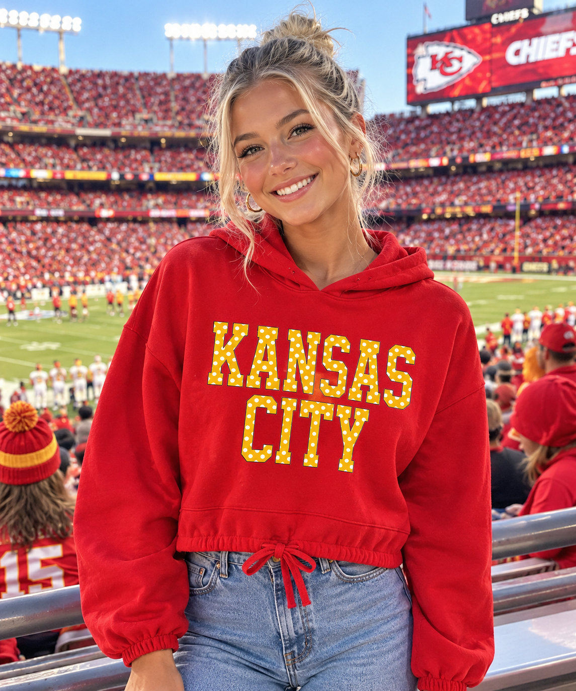A woman wearing a red cropped hoodie with 'KANSAS CITY' in yellow polka dot block letters standing in a crowded football stadium.