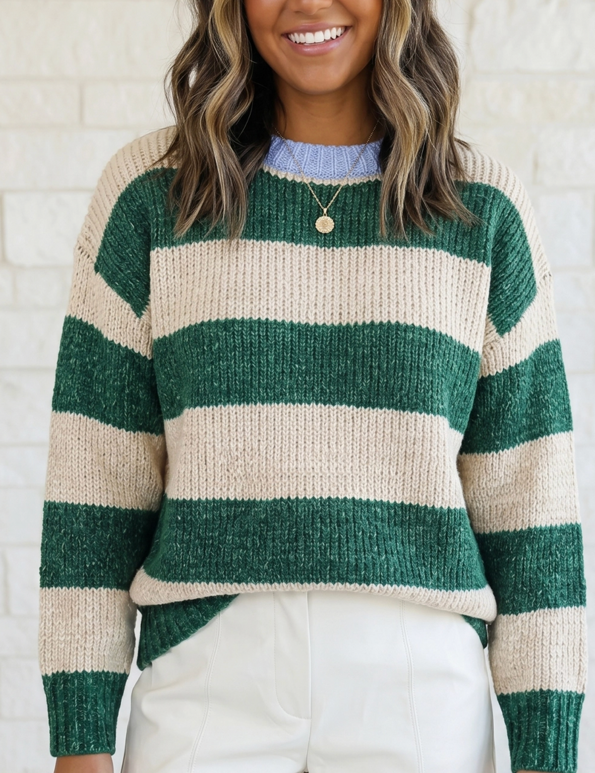 A woman wearing a green and cream striped knit sweater with white shorts, standing against a white brick wall.