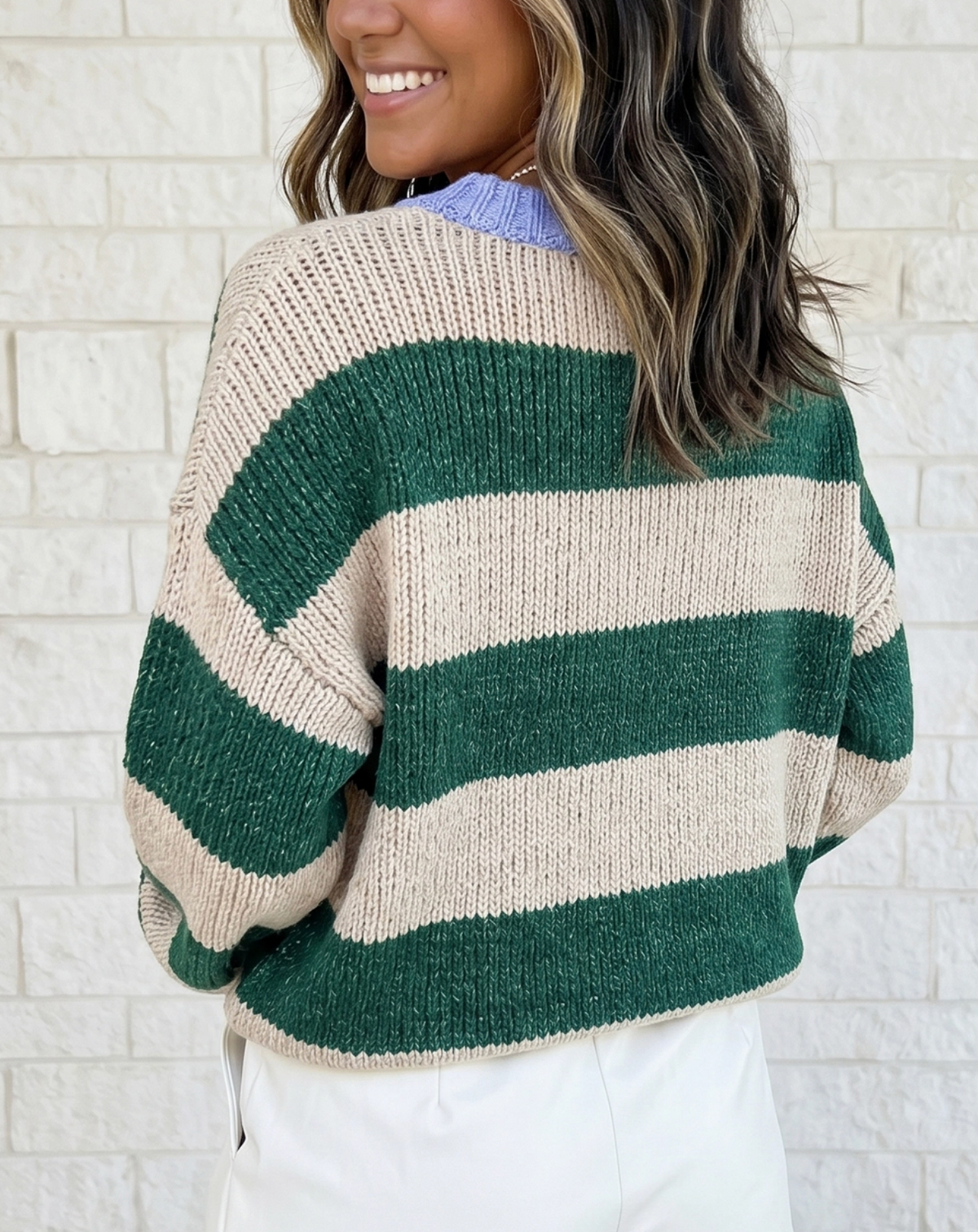 A woman wearing a chunky knit sweater with wide horizontal stripes in forest green and cream, shown from the back against a white brick wall.