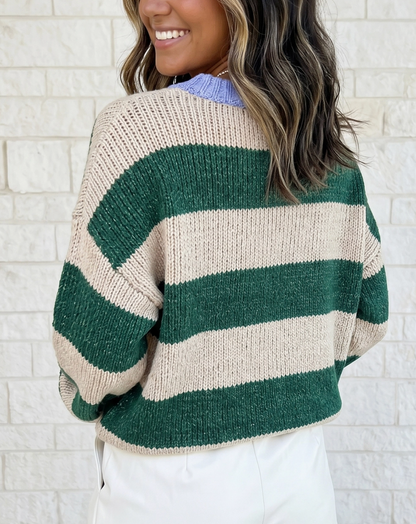 A woman wearing a chunky knit sweater with wide horizontal stripes in forest green and cream, shown from the back against a white brick wall.