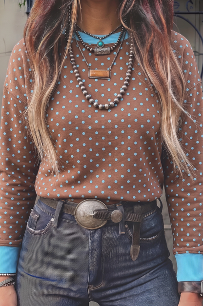 A person wearing a brown long-sleeve top with light blue polka dots, layered with multiple necklaces, and dark blue jeans with a large silver belt buckle.