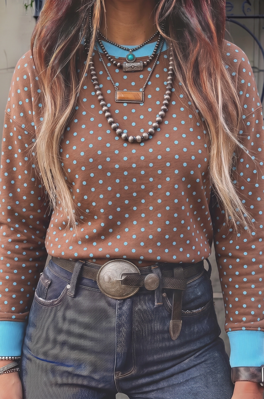 A person wearing a brown long-sleeve top with light blue polka dots, layered with multiple necklaces, and dark blue jeans with a large silver belt buckle.